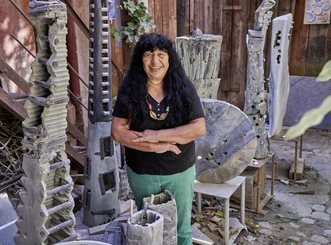 Künstlerin Claudia Endres im Innenhof ihres Ateliers in Nürnberg-Gostenhof. Fotoaufnahme von Ralph Schirrmeister, © VG Bild-Kunst, Bonn 2026. Die Fotoaufnahme zeigt die Künstlerin Claudia Endres im Innenhof ihres Ateliers in Nürnberg-Gostenhof. Sie ist eine Frau mit langen schwarzen Haaren, trägt ein schwarzes Shirt und eine grüne Hose, und lächelt freundlich mit überkreuzten Armen in die Kamera. Im Hintergrund ist eine große Zahl ihrer Kunstwerke aus Stein zu sehen, die sie wie kleine Türme umringen. Die Fotoaufnahme stammt von Ralph Schirrmeister.