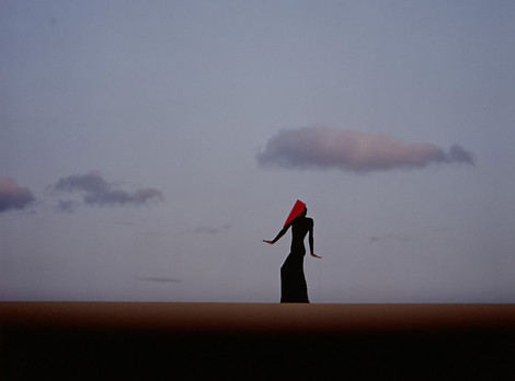 Lajos Keresztes: Aus der Serie „Licht Zeichen Sprache“, 0014, 1985; © Lajos Keresztes 2025. Eine einzelne Person mit langem Kleid und rotem Tuch über dem Kopf steht aufrecht in weiter Landschaft. Der Himmel ist klar und hell, mit wenigen, locker verteilten Wolken. Die Szene wirkt minimalistisch, der Horizont verläuft gerade und teilt das Bild in zwei Flächen.