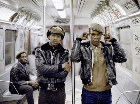 Jamel Shabazz: The Righteous Brothers, NYC 1981 © Jamel Shabazz, courtesy Galerie Bene Taschen, Köln. Die Abbildung zeigt eine Fotografie von Jamel Shabazz aus dem Jahr 1981. Zwei junge afroamerikanische Männer mit dunklen Lederjacken und Schirmmützen stehen in einem New Yorker U-Bahn-Wagen. Einer verschränkt die Arme, der andere hält sich die Zeigefinger an den Kopf. Im Hintergrund sitzt ein weiterer afroamerikanischer Mann auf einer Bank. Die Wände und Türen des Wagens sind mit Graffiti bedeckt.