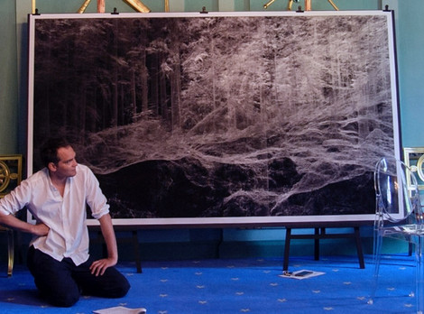 Simon Strauß vor dem Werk ‚Ammergebirge Bayern 1‘ von Ralph Mecke. Fotoaufnahme von Ruth Walz. Die Abbildung zeigt eine Fotografie eines Mannes, der vor einem großen Kunstwerk des Künstlers Ralph Mecke kniet. Bei dem Mann handelt es sich um Schriftsteller Simon Strauß, die Fotoafnahme stammt von Ruth Walz.