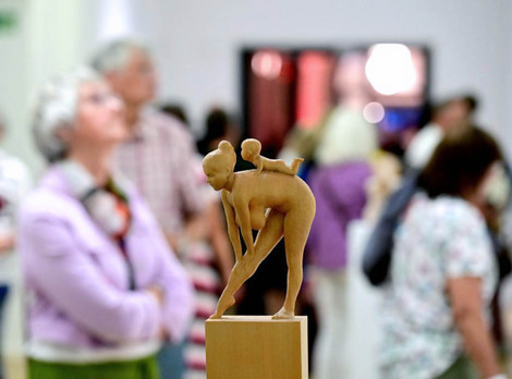 Besucher*innen bei der Eröffnung der Ausstellung zum NN-Kunstpreis 2025 | Fotoaufnahme von Joachim Winkler (NN) Die Abbildung zeigt im Vordergrund eine kleine Holzskulptur. Es handelt sich um eine sich bückende nackte Frau, auf deren Rücken ein Baby krabbelt. Im Hintergrund sind Personen bei der Betrachtung von anderen Kunstwerken zu sehen. Die Fotoaufnahme stammt von Joachim Winkler.